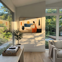 She sits within the white architecture, watching a sail move across the gold-lit sea. The orange drapery pulls the sunset into the room, making longing feel warm and exact.