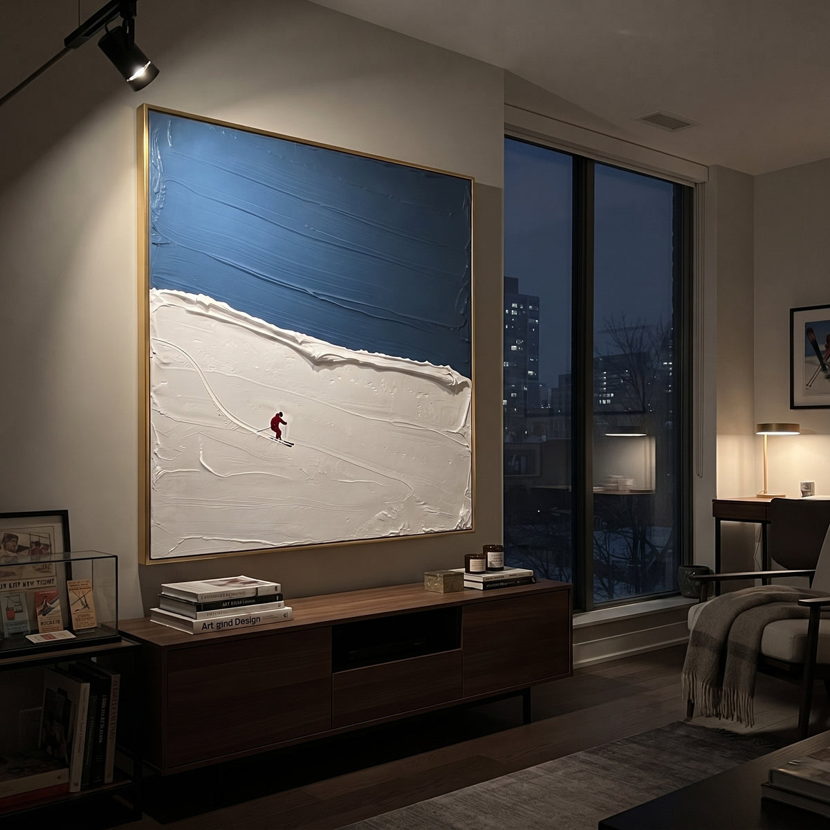 Blue Ridge Solo Skier focuses on one red skier crossing a quiet blue ridge below a deep winter sky. The artwork shows a winter ski scene built from the actual image: visible downhill figures, curved snow tracks, raised ridgelines, and alpine space arranged in a balanced composition. The blue, white, red palette gives the piece a cold mountain atmosphere while the thick impasto surface creates shadows along each snowbank, ski line, and textured ridge. This is presented as hand-painted textured